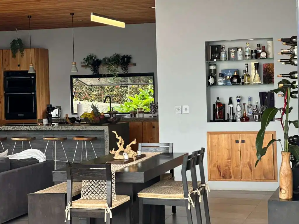 Interior de hermosa casa campestre moderna con cocina, comedor y barra de bar, rodeada de exuberante vegetación.