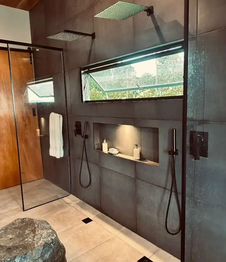 Moderna ducha de baño en casa campestre con dos regaderas tipo lluvia, azulejos grises y vista a la naturaleza.