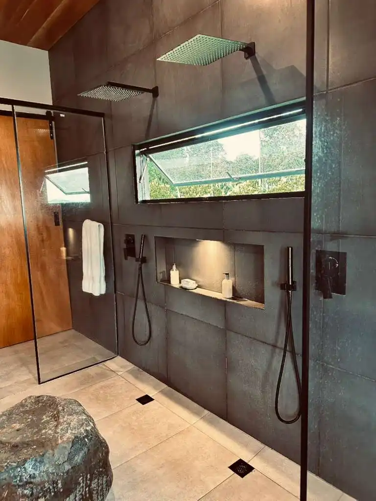 Moderna ducha de baño en casa campestre con dos regaderas tipo lluvia, azulejos grises y vista a la naturaleza.