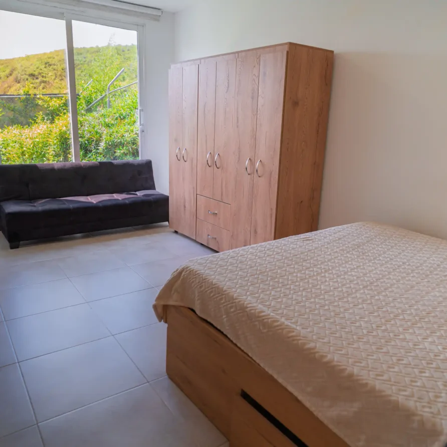 Dormitorio luminoso con cama, sofá, armario de madera y ventana con vista a la vegetación, ideal para cabaña Lago Calima.