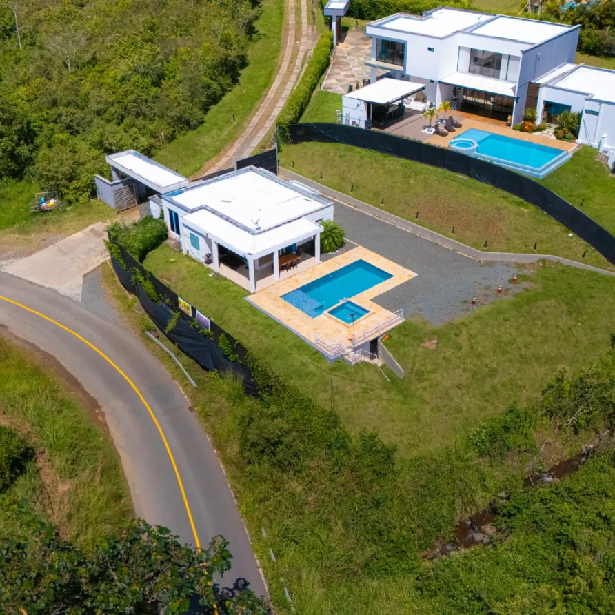 Vista aérea de dos casas modernas con piscinas, rodeadas de exuberante vegetación y una carretera curva.