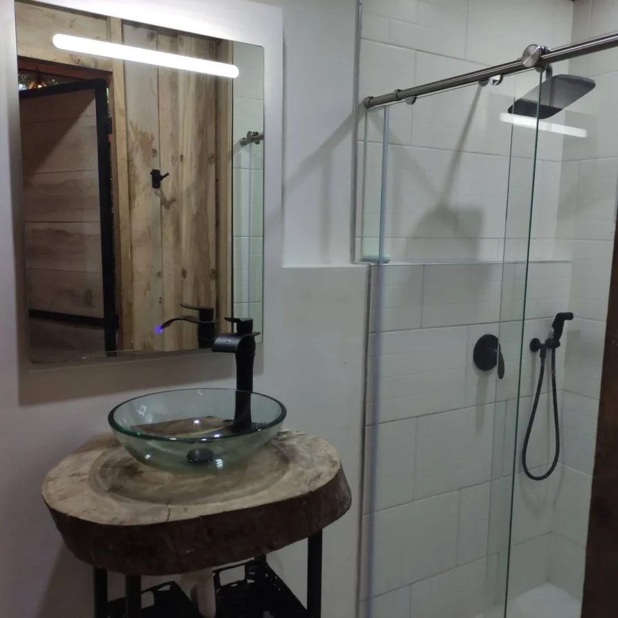 Planes Turisticos 116 Baño moderno con lavamanos de cristal sobre tronco de madera rústico, espejo LED y ducha elegante con grifería negra.