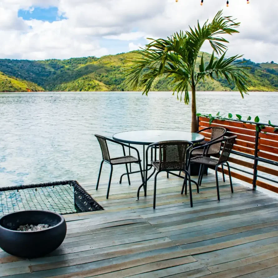 Terraza de glamping flotante sobre el Lago Calima, con mesa, sillas, palmera y BBQ, ideal para disfrutar de la vista.